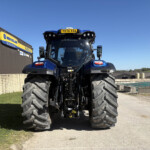 Front view of a blue New Holland tractor with oversized tires, parked beside a building with a yellow New Holland sign.