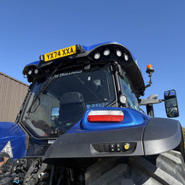 Blue New Holland tractor cabin with yellow license plate reading YX74 XXA and orange beacon on top against a clear sky.