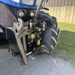 Close-up of the front of a blue tractor showing two round headlights and a hitch assembly, with a brick wall and grass in the background.