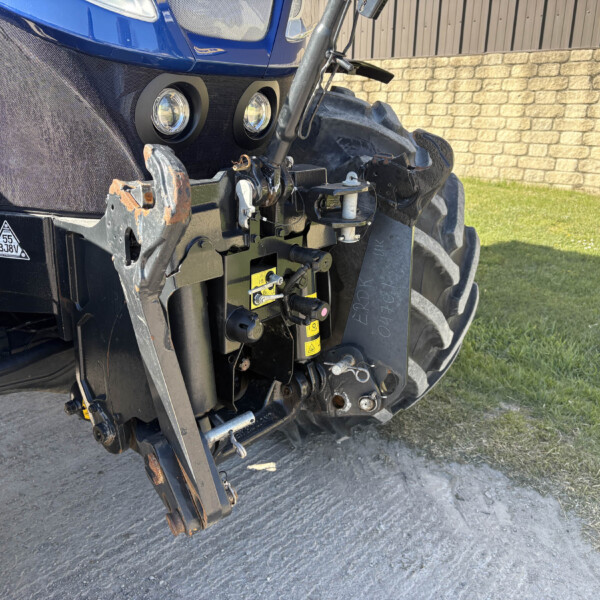 Close-up of the front of a blue tractor showing two round headlights and a hitch assembly, with a brick wall and grass in the background.