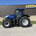 Blue New Holland tractor seen from the front beside a dark corrugated wall with a large yellow and blue "New Holland Agriculture" sign above.