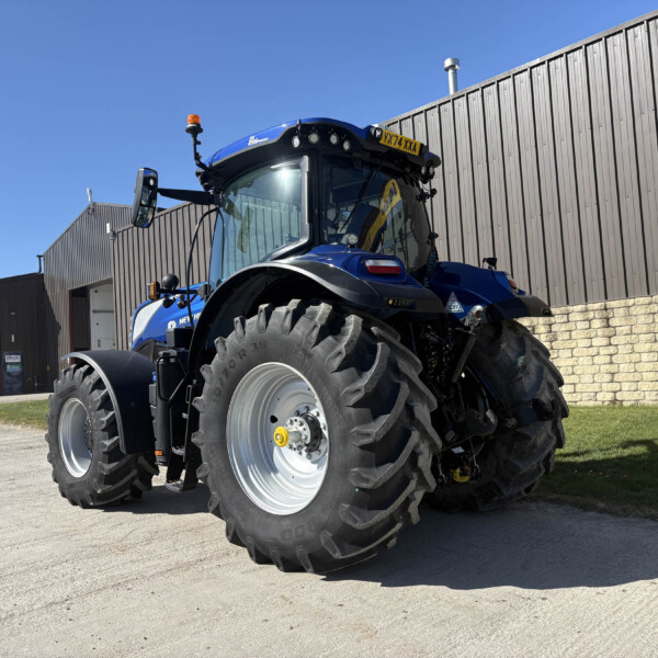 Blue agricultural tractor with oversized tires parked on a concrete lot beside a corrugated metal building under a clear blue sky.