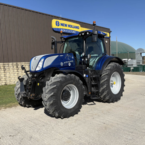 Blue New Holland tractor with oversized tires parked in front of a brown industrial building under a clear blue sky (advertising sign reads New Holland Agriculture).