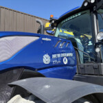 Close-up of a blue New Holland tractor front, showing the hood, grille, and windshield with dealership stickers under a clear sky.