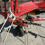 Close-up of red industrial machinery frame with hoses, cables, and a wheel on a concrete surface.
