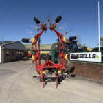 Industrial agricultural rake/harvester with multiple tines in a red-yellow frame, displayed in a lot outside a farm equipment dealership under a clear blue sky.