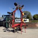 Red agricultural implement with multiple tines and wheels on display in a parking lot, with a blue tractor and a brick wall nearby against a clear sky.