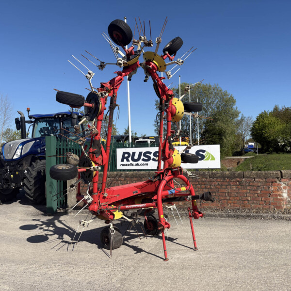Red agricultural implement with multiple tines and wheels on display in a parking lot, with a blue tractor and a brick wall nearby against a clear sky.