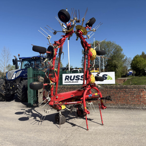 Red farming implement with many tines attached to a tractor in a parking lot, with a blue tractor in the background under a clear blue sky.