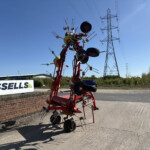 Red agricultural equipment sculpture with multiple arms and wheels beside a brick wall and Russell's sign, under a clear blue sky.