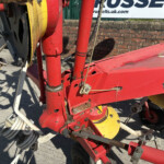 Close-up of red agricultural machinery with coiled hoses and metal components on a concrete surface, against a brick wall and banner in the background.