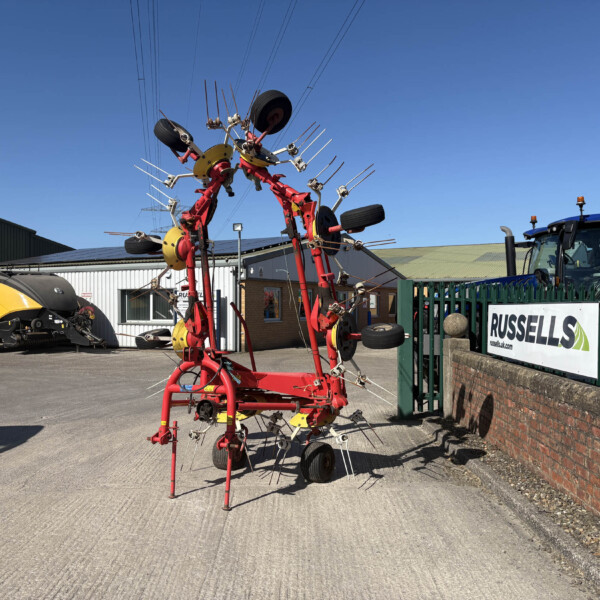 Red multi-axle farming implement with numerous tines standing in a lot beside tractors and a brick wall under a clear blue sky