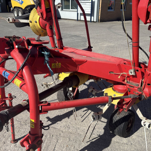 Red agricultural implement with yellow discs and exposed hoses/ropes on a concrete lot; close view of machinery components with a building in the background.