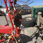 Red agricultural machinery with tangled metal rods and a large worn tire in a yard near a brick wall and green fence.