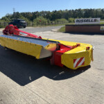 Close-up of a large yellow and red industrial roadwork mower or barrier attachment on a trailer, with a brick wall and Russell's sign in the background.