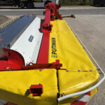 Red hydraulic machine arms mounted on a trailer, with a bright yellow safety mat along the side by a road.