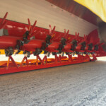 Close-up of a red farming implement with multiple red tines and clamps mounted under a machine on a paved surface.