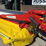 Red agricultural mower attachment with a yellow protective guard, Pöttinger logo visible, outdoors at a display area.