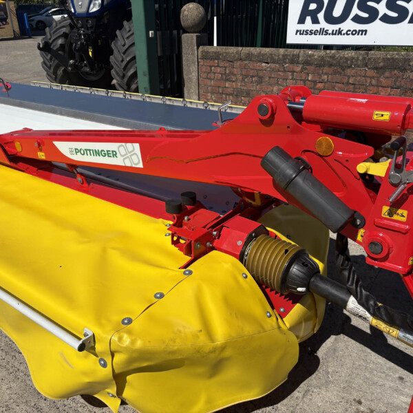 Red agricultural mower attachment with a yellow protective guard, Pöttinger logo visible, outdoors at a display area.