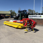 Yellow agricultural mower attachment in the foreground at a farm equipment yard, with a blue tractor and Russell's sign visible in the background under a clear blue sky.