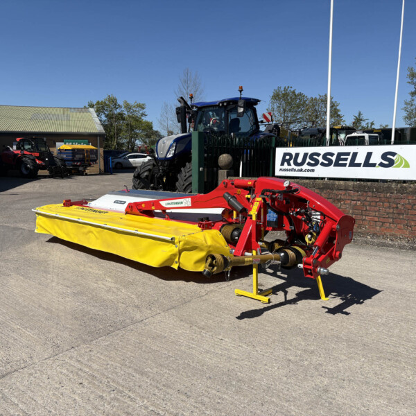 Yellow agricultural mower attachment in the foreground at a farm equipment yard, with a blue tractor and Russell's sign visible in the background under a clear blue sky.