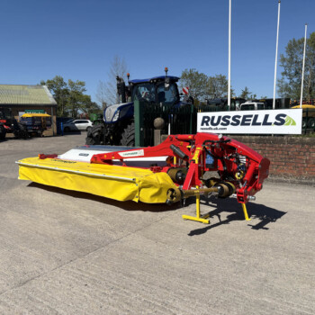 Outdoor lot with farm equipment: a large blue tractor behind a green fence, sign reading 'RUSSELLS' in the foreground, and red and yellow machinery at the front on a sunny day.