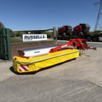 Wide view of an industrial yard with a bright yellow safety barrier in the foreground, a brick wall, and a Russell's sign beside a loading trailer and trucks under a blue sky.