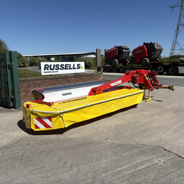 Wide view of an industrial yard with a bright yellow safety barrier in the foreground, a brick wall, and a Russell's sign beside a loading trailer and trucks under a blue sky.
