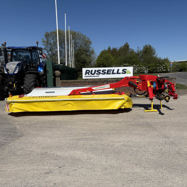 Blue agricultural tractor in a yard with stacked farming equipment and a Russell's sign against a bright blue sky+green trees in background.