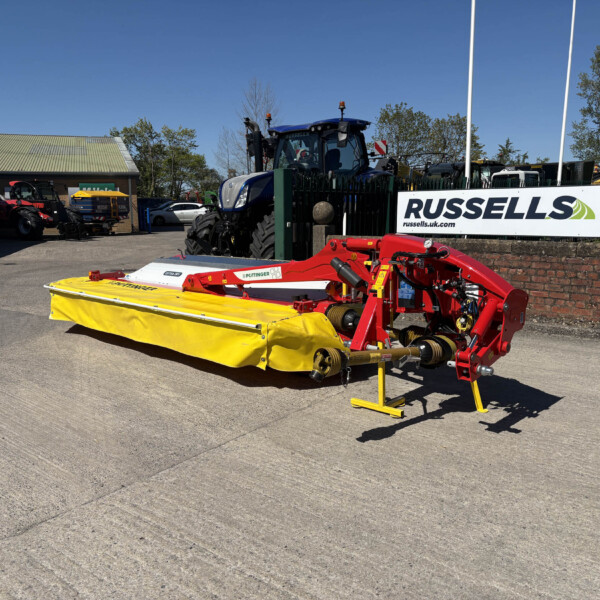 Blue farm tractor behind a green fence with a large yellow mower in the foreground, outdoor equipment lot under a clear blue sky, Russell's sign visible on the right.