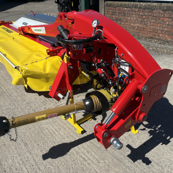 Red agricultural machine with exposed hydraulic hoses and a yellow protective cover, parked on a paved surface near a brick wall; gauge and metal components visible.