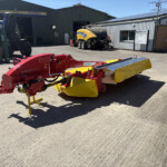 Red agricultural mower attachment in the foreground at a farm equipment yard, with a blue-and-yellow building and large machinery in the background.