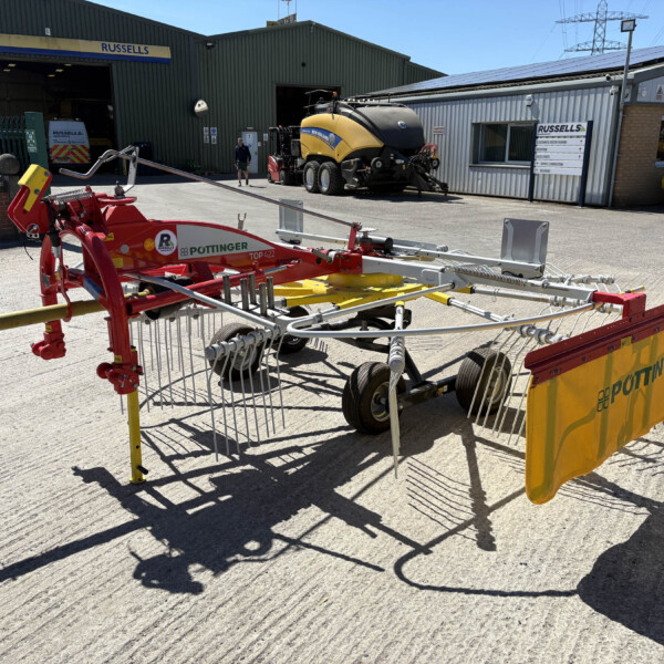 Agricultural machinery in a factory yard, with a red Pöttinger implement in the foreground and a yellow and black baler behind, near a green warehouse labeled Russell's Agriculture.