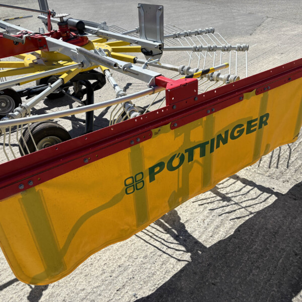Close-up of a red and yellow agricultural implement with metal tines and gears, resting on a concrete surface; the banner shows the word PÖTTINGER.