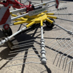 Agricultural rake with a bright yellow central hub and radiating metal tines, labeled Pöttinger, resting on a concrete surface.