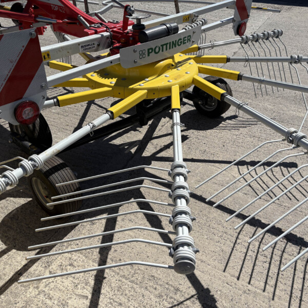 Agricultural rake with a bright yellow central hub and radiating metal tines, labeled Pöttinger, resting on a concrete surface.