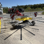 Red and yellow Pöttinger rake on a paved road with a white Russell's truck in the background and trees beyond.
