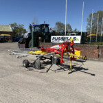 Agricultural equipment lot with a blue tractor behind a green fence and various red and yellow farming implements in front; sign reads 'RUSSELLS' in background.
