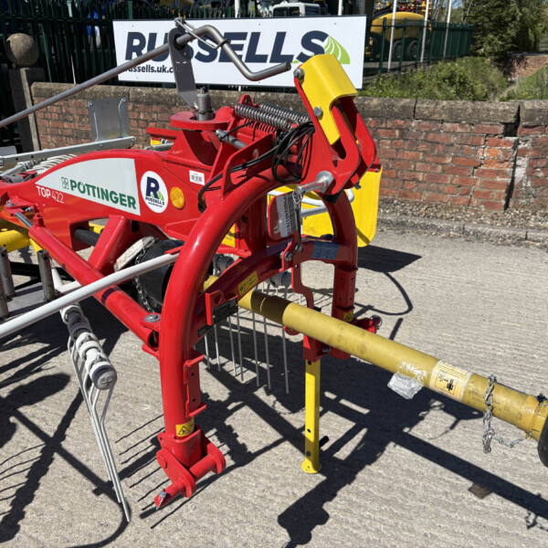 Close-up of a red farming implement with yellow guard and hydraulic hoses, outdoors beside a brick wall and fence, during the day.