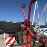 Close-up of red agricultural or construction machinery frame with a large black multi‑part wheel, safety barriers, and a clear blue sky in the background, outdoors at a yard.