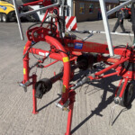 Red agricultural machine frame with hoses, cables, and warning stickers outdoors in a yard. A yellow vehicle and a person are visible in the background.