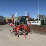 Red agricultural equipment with long tines parked in a lot beside a brick building under a clear blue sky