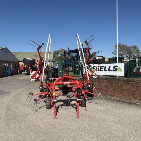 Red agricultural equipment with long tines parked in a lot beside a brick building under a clear blue sky