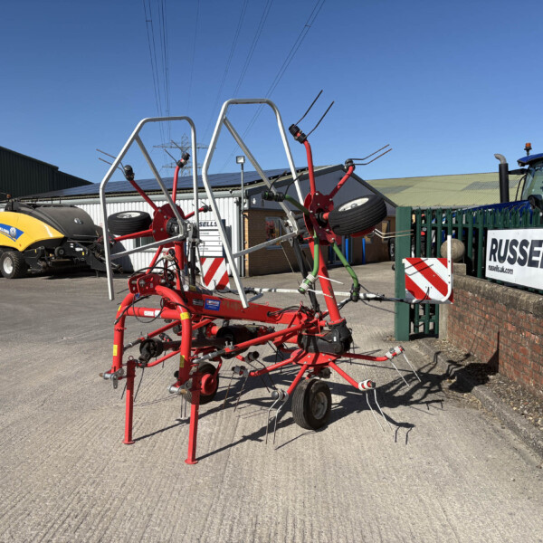 Red agricultural implement in the foreground with a yellow New Holland mower on the left and a blue tractor behind a green fence under a clear blue sky.