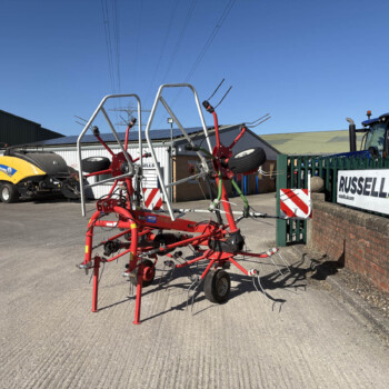 Red agricultural implement with metal Y-shaped arms in foreground, parked in a lot beside a green fence and Russell's sign in the background
