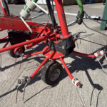 Close-up of a red agricultural implement with a metal frame, wheels, and tangled hoses on a concrete surface.