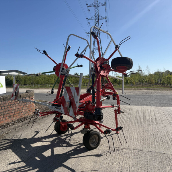 Red farming implement with multiple tines and wheels, parked beside a brick wall on a concrete lot under a clear blue sky