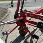 Close-up of red industrial machinery with springs, cables, and a wheel on a paved surface beside a brick wall.