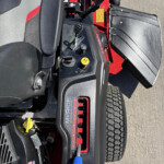 Close view of a vehicle operator station (tractor) showing a black armrest, control levers, yellow hydraulic cap, and red body panels with tires in the background