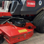 Close-up of a Toro ride-on lawn mower deck: red cutting deck with yellow warning labels, black engine housing, seat with red stitching, and a large rear tire visible.